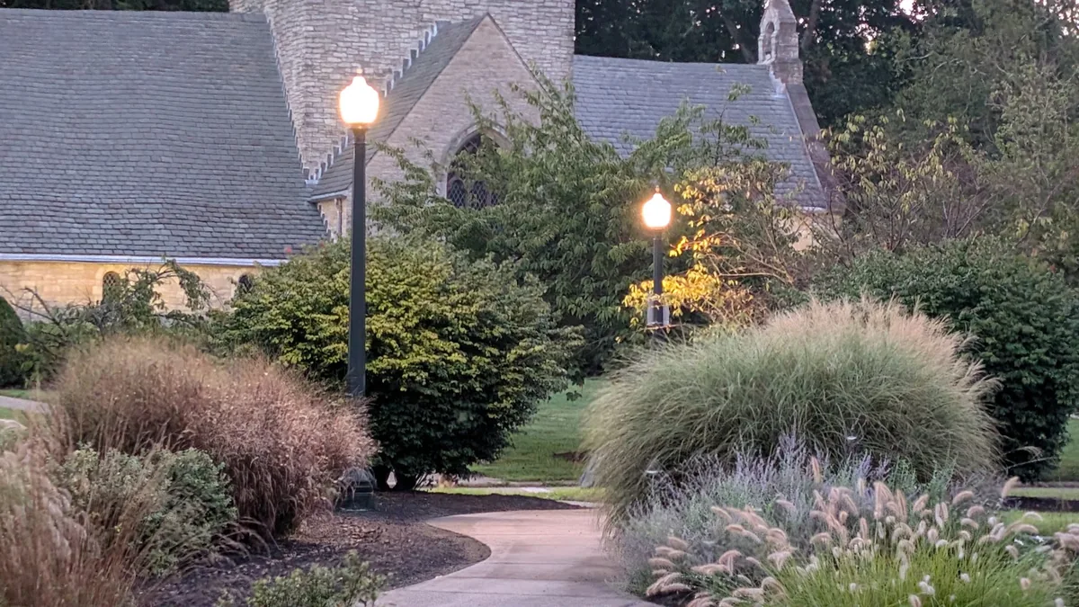 Decorative LED post top lanterns lit at dusk along a village path, with a stone church and planted borders behind